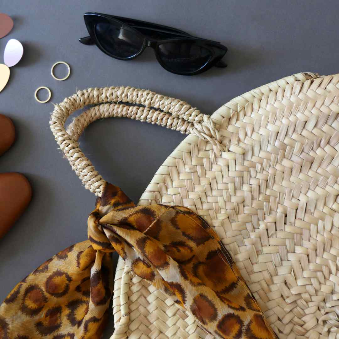 Partial image of a round raffia beach bag with a leopard print scarf tied to the handle and a pair of sunglasses and small gold hoop earrings