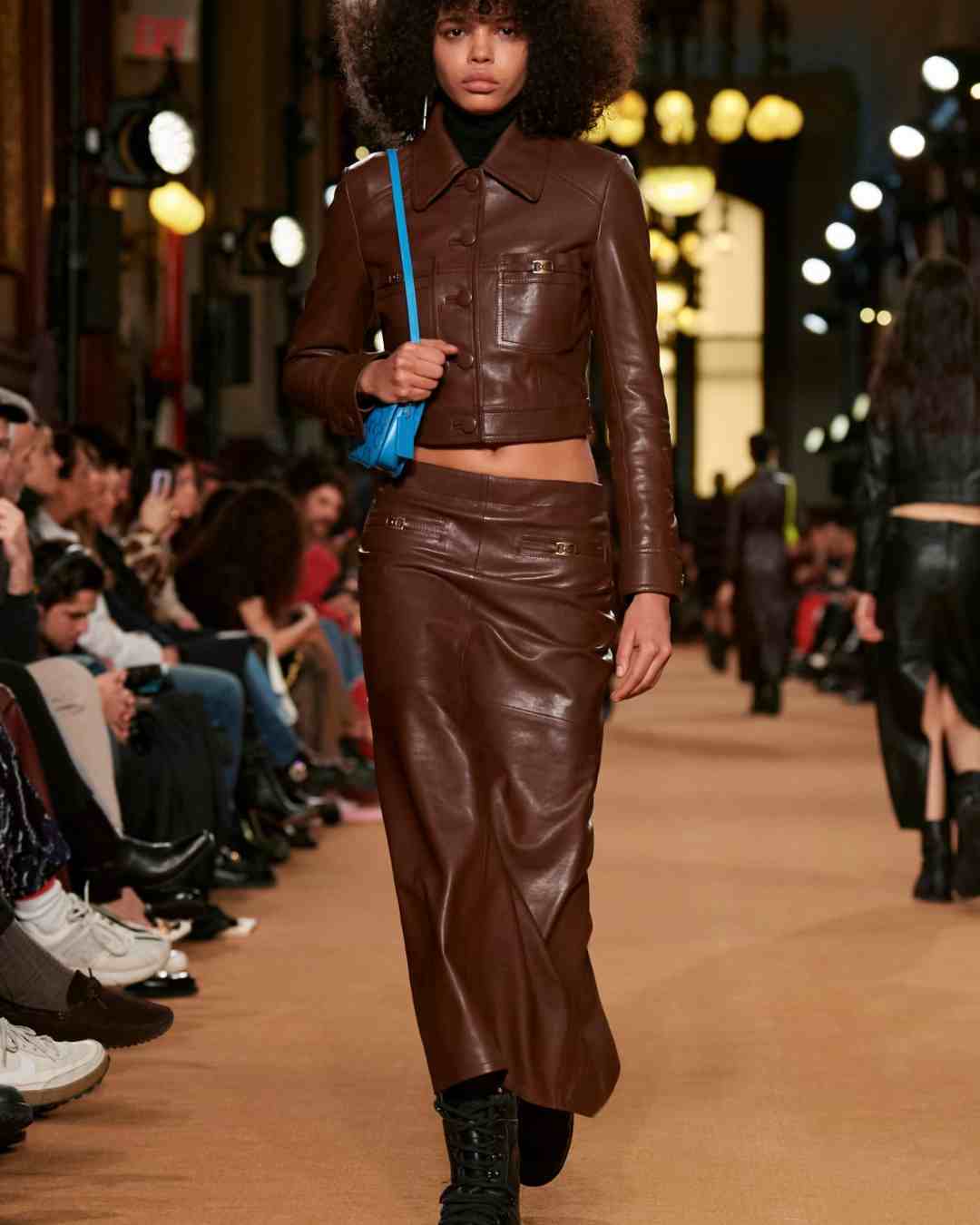 Women wears cholcolate brown leather jacket buttoned up and cropped at the waist and a leather maxi skirt in the same colour with boots and carries and aqua blue small bag over the shoulder